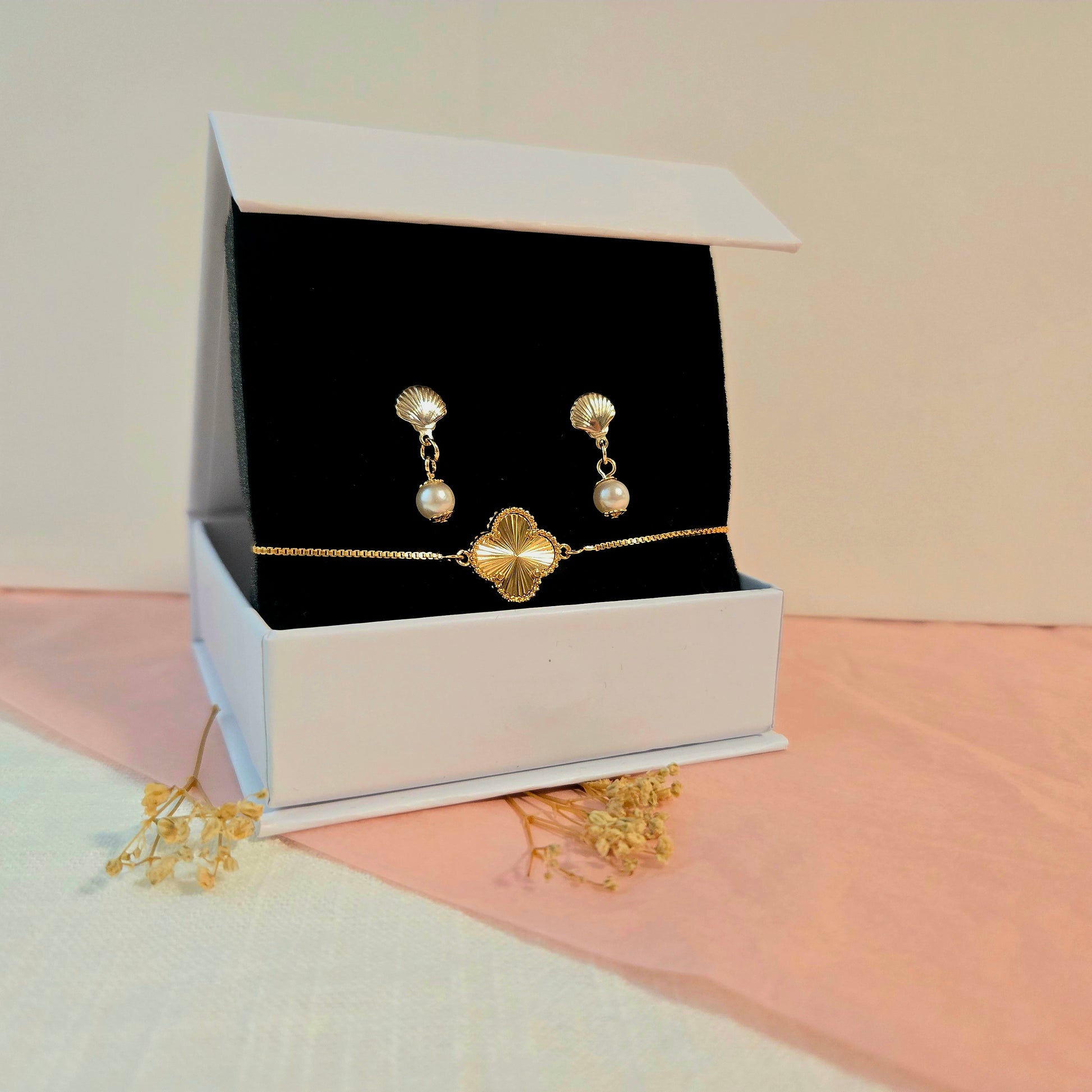 Matching gold charm bracelet and pearl drop earrings displayed in a white jewelry box with black velvet insert, styled on blush pink fabric and dried florals