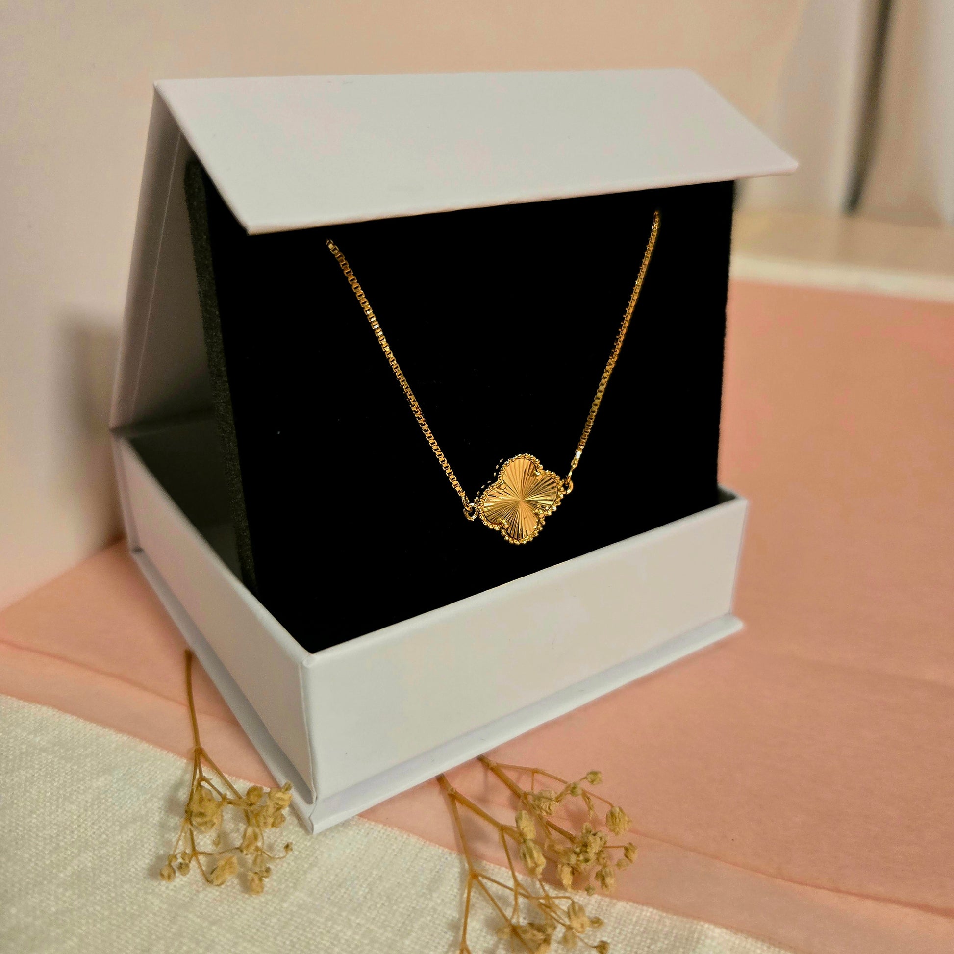 Gold butterfly charm bracelet displayed in a white jewelry box with black velvet interior, styled on blush pink fabric with dried florals