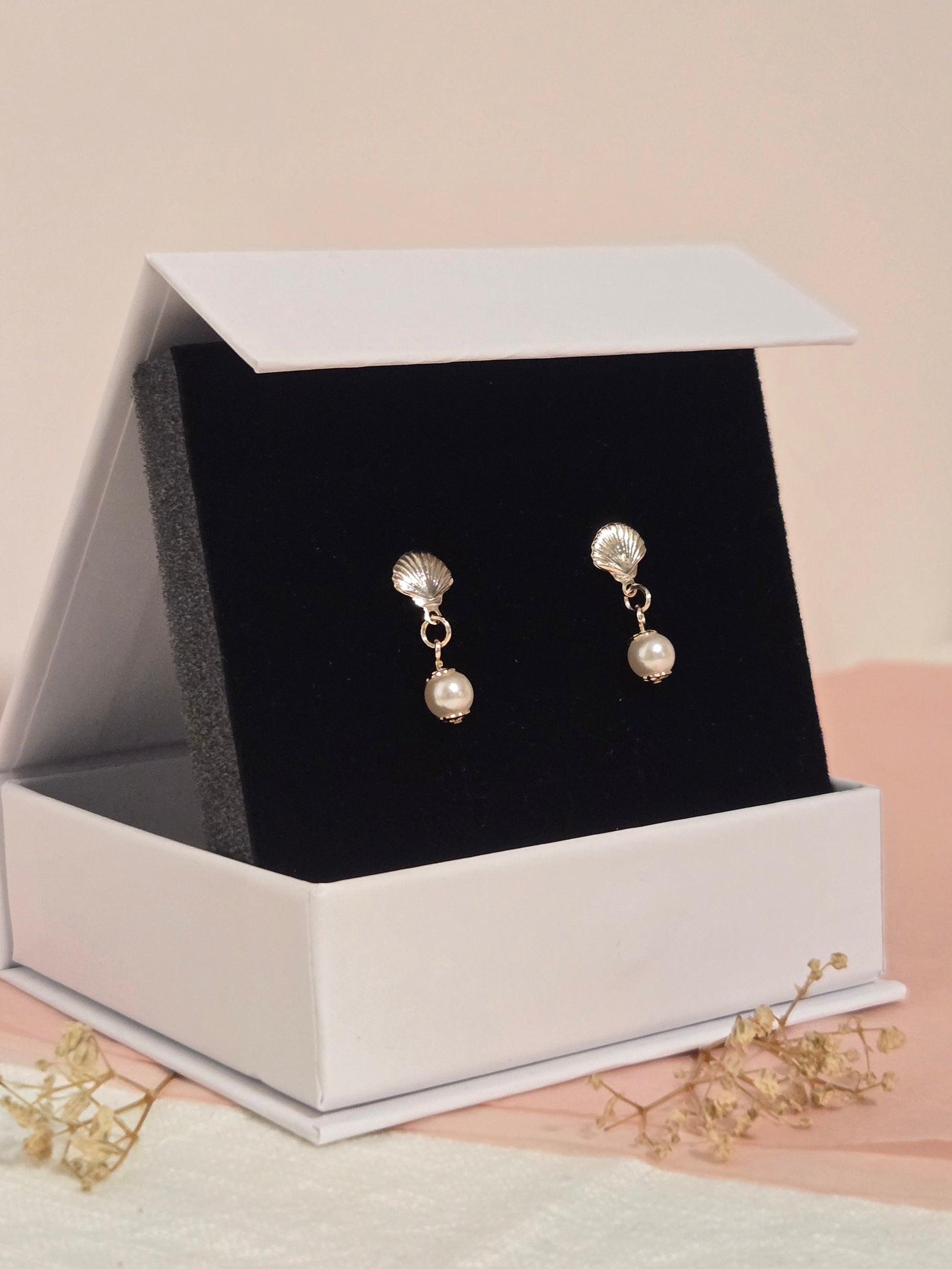 Gold seashell and pearl drop earrings displayed in a white jewelry box with black velvet lining, styled on blush and cream fabric with dried florals