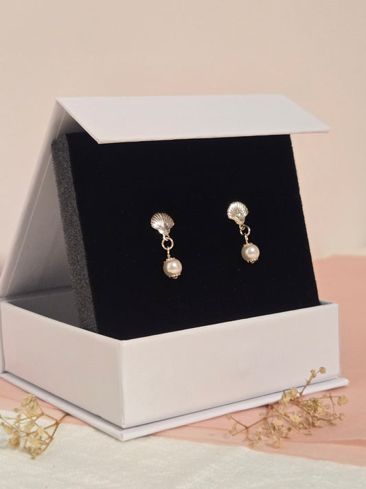 Gold seashell and pearl drop earrings displayed in a white jewelry box with black velvet lining, styled on blush and cream fabric with dried florals